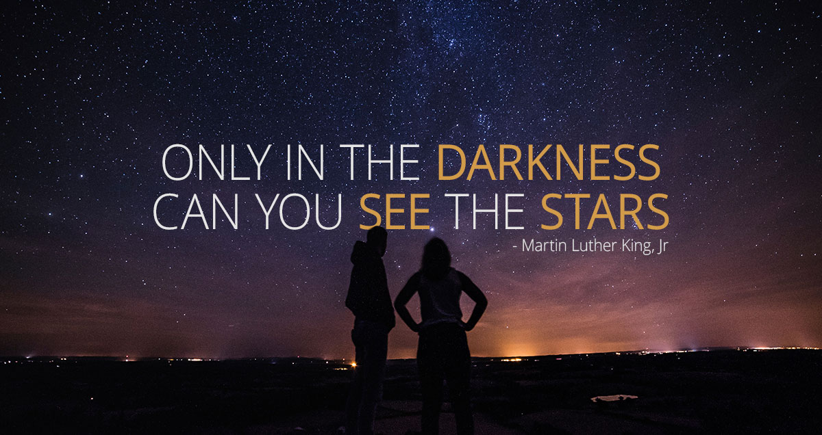 A couple stands together, looking at the night sky with a quote from Marin Luther King, Jr. "Only in the darkness can you see the stars."